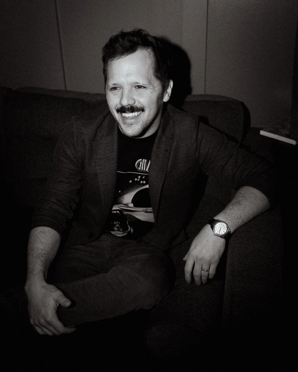 Black-and-white portrait of Nick Rowley, smiling, seated in an armchair wearing a blazer over a graphic t-shirt.